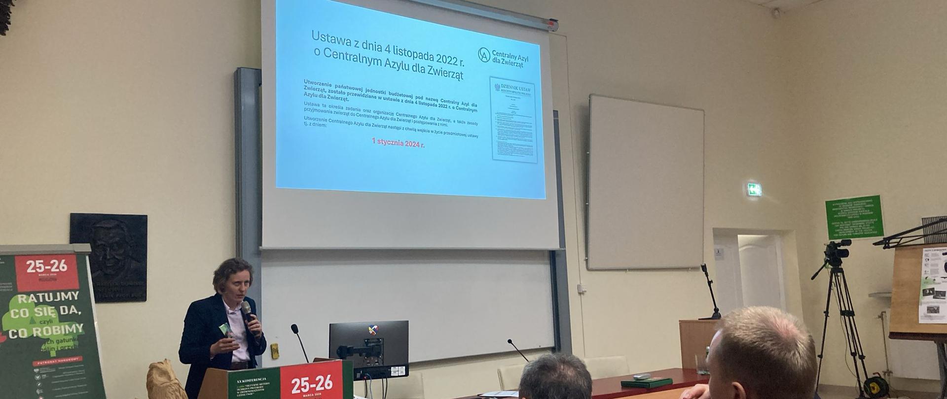 Sala konferencyjna w Rogowie podczas prezentacji multimedialnej dotyczącej ochrony zagrożonych gatunków; widoczni uczestnicy konferencji oraz ekran z treścią wystąpienia.