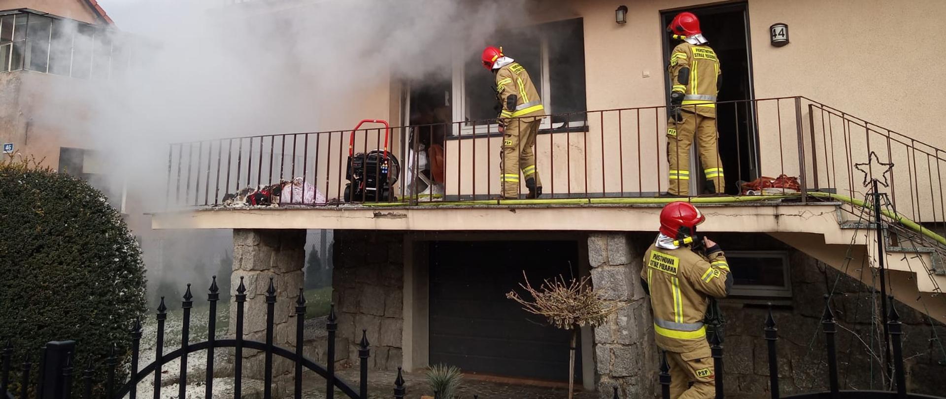 Strażacy przygotowują się do akcji gaśniczej. W tle budynek jednorodzinny z którego wydobywają się kłęby dymu.