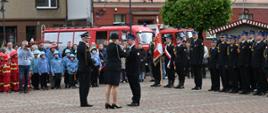 Strażak dekorowany na środku placu. Medal przypina pani w mundurze. obok niej stoi jeszce strażak. Za nimi w tle inni strażacy zgromadzeni na zbiórce. nad. ich głowami zielone drzewo i ściana budynku. 