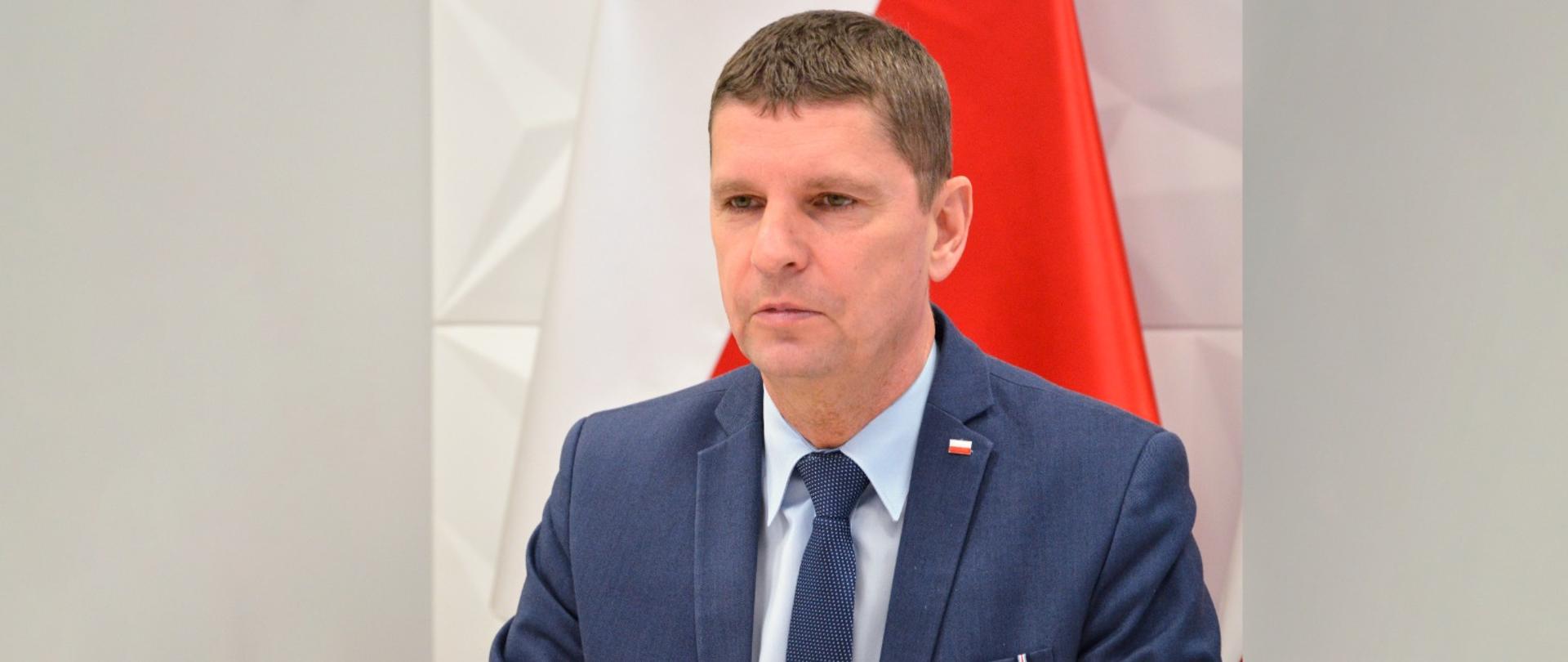 Za brązowym stołem siedzi wiceminister Piontkowski, za nim na żółtej ścianie wisi polska flaga.