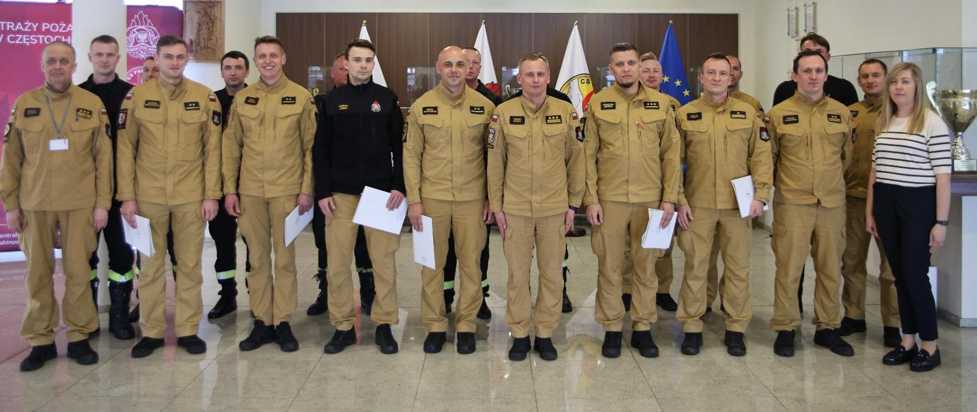 Pamiątkowe zdjęcie grupowe funkcjonariuszy PSP będących uczestnikami szkolenia "Prąd elektryczny, a zagrożenia dla strażaków" (15 osób) w towarzystwie Zastępcy Komendanta Centralnej Szkoły PSP, kobiety oraz trzech funkcjonariuszy CS PSP