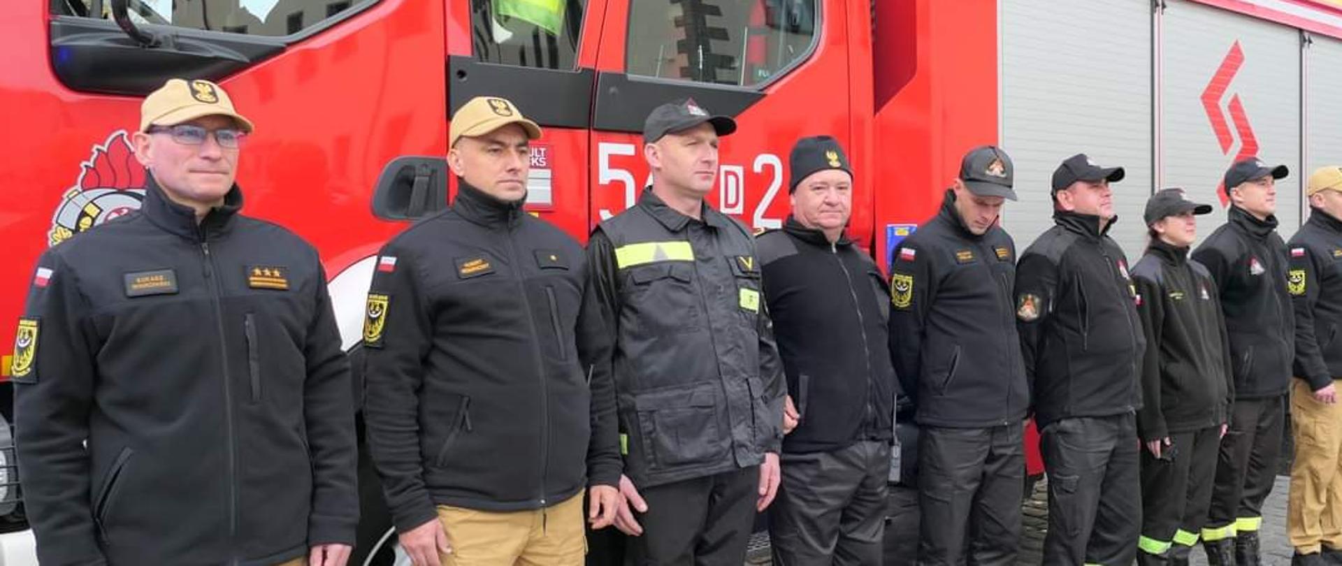 Strażacy stojący na baczność na tle samochodu pożarniczego.