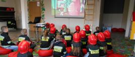 Dzieci na sali ognik siedząc na poduszkach oglądają edukacyjną bajkę wyświetlaną na rzutniku. 