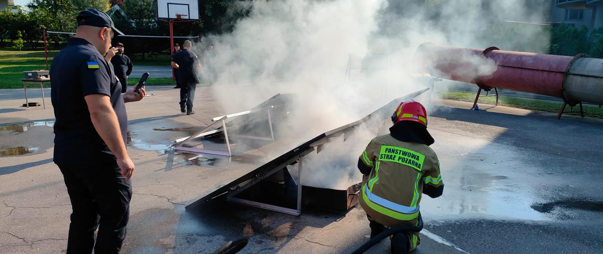 Na placu szkoleniowym gaszony jest panel fotowoltaiczny, widać strażaka oraz ludzi w mundurach z flagą Ukrainy