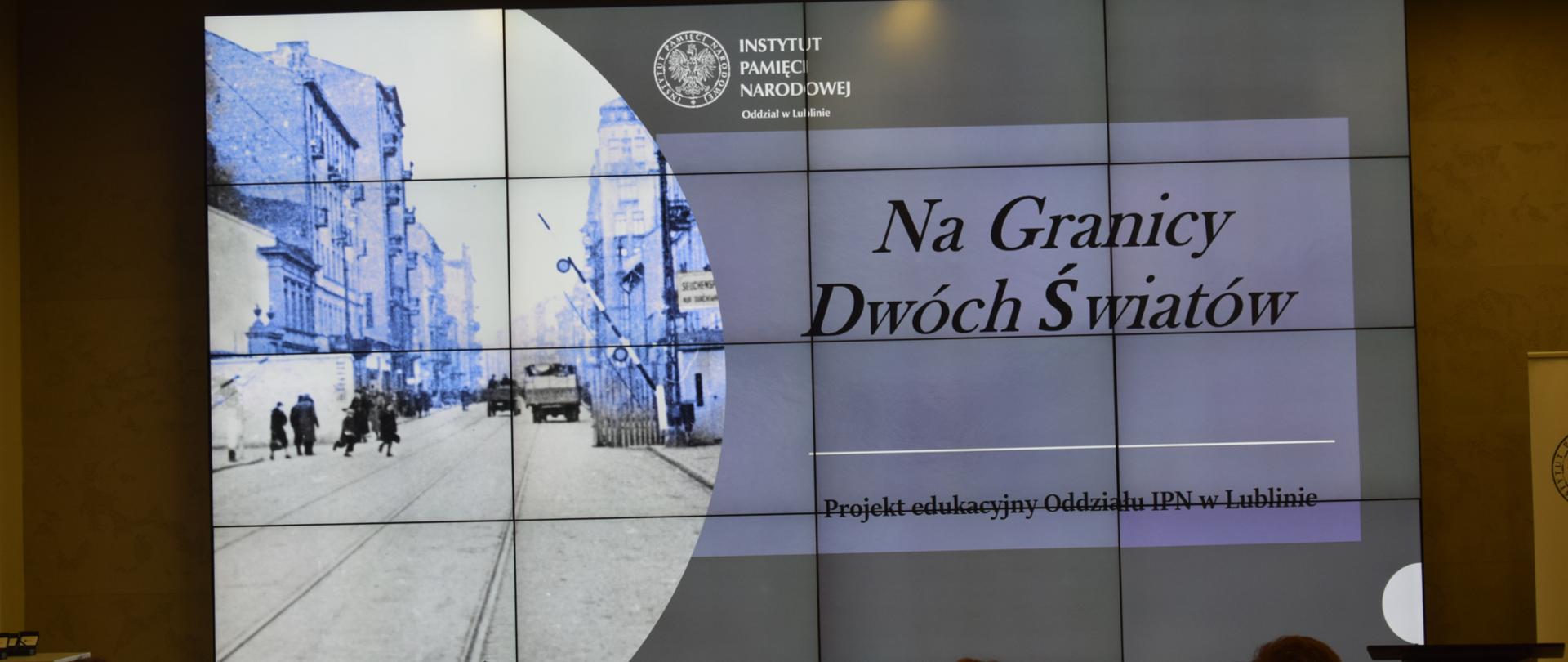 Grupa uczniów w sali IPN w Lublinie ogląda slajd przedstawiający monochromatyczną fotografię na której widać ludzi i dwa samochody ciężarowe na tle zabudowań miejskich okresu międzywojennego, a także napis "Na granicy dwóch światów", "Projekt edukacyjny Oddziału IPN w Lublinie", "Instytut Pamięci Narodowej Oddział w Lublinie".