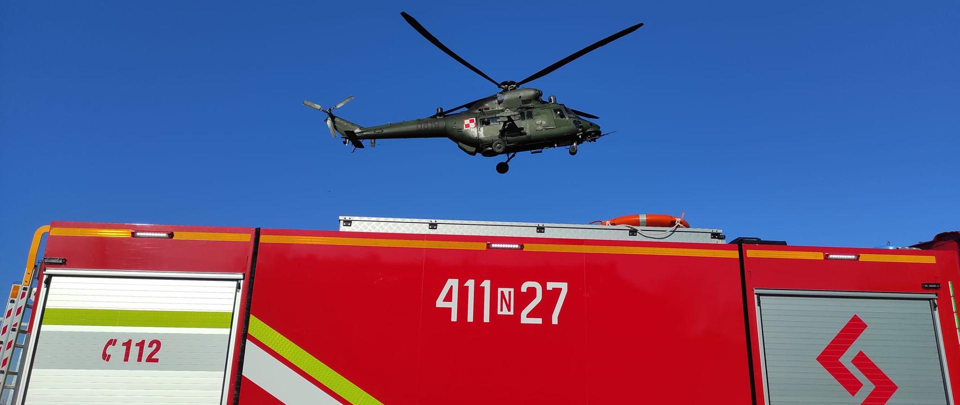 Nad czerwonym samochodem strażackim unosi się wojskowy śmigłowiec. Niebieskie niebo.