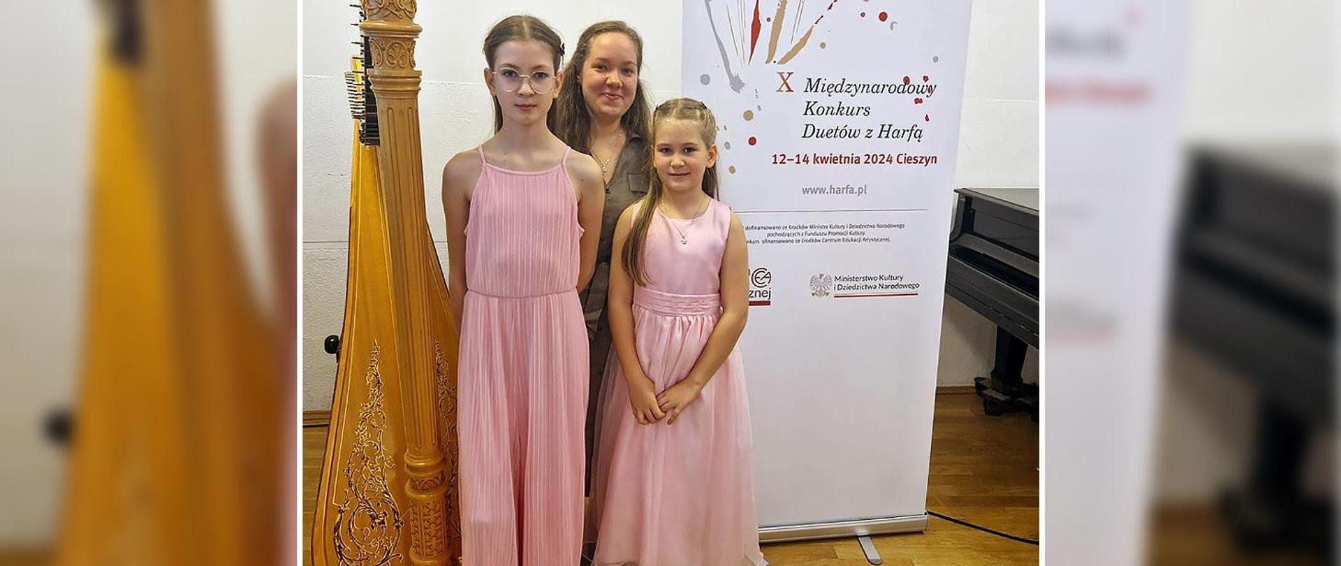 Nauczycielka i dwie uczennice stoją w auli, obok nich stoi harfa, za nimi baner z tytułem i datą konkursu.