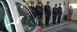 Przekazanie samochodu w OSP Jutrosin. Garaż w remizie OSP Jutrosin. Na pierwszym planie fragment przekazywanego samochodu, a w tle druhowie OSP uczestniczący w zbiórce. 