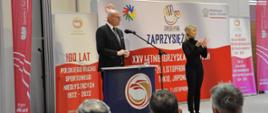 Podsekretarz stanu w Ministerstwie Sportu i Turystyki Ireneusz Nalazek przemawia zaprzysiężeniu reprezentacji Polski na XXV Letnie Igrzyska Głuchych - Tokio 2025. Obok niego stoi tłumaczka języka migowego. Za jego plecami okolicznościowe ścianki w biało-czerwonych barwach. Z boku czerwony winder z logotypem MSiT.