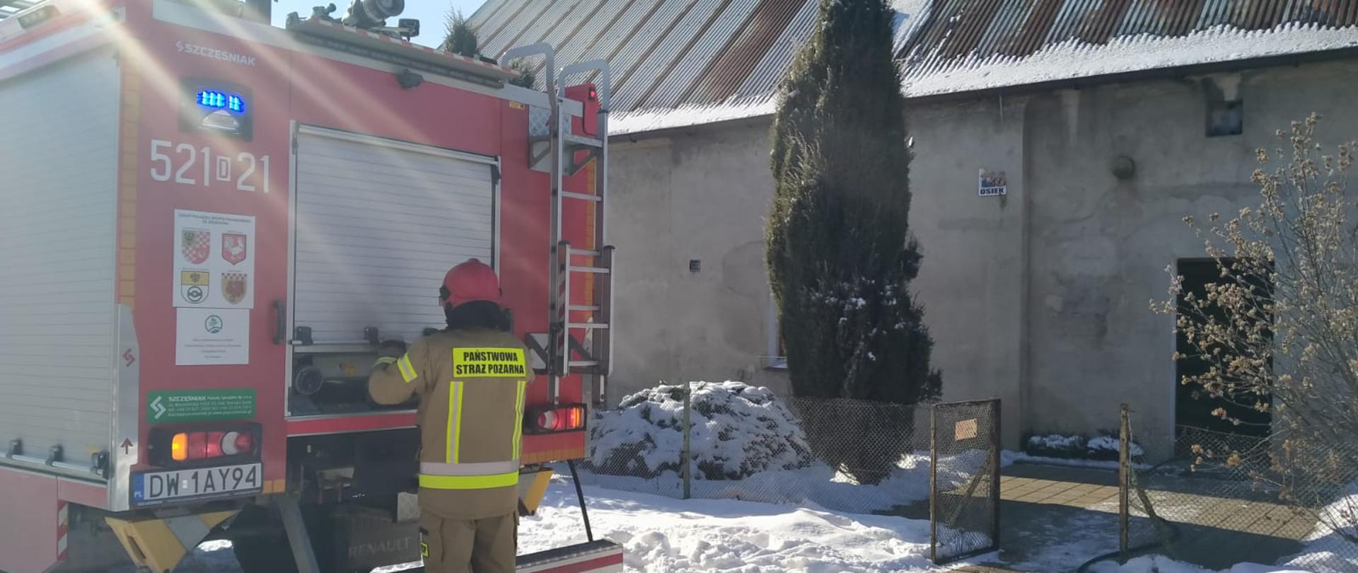 Na zdjęciu widać tył samochodu strażackiego oraz strażaka, który obsługuje autopompę. Waż "szybkiego natarcia" rozwinięty jest do wnętrza budynku mieszkalnego, po prawej stronie.
