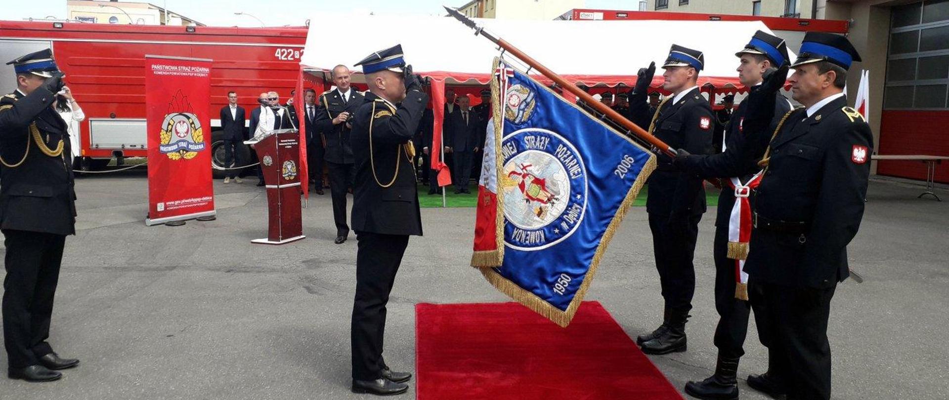 Na zdjęciu komendant powiatowy Państwowej Straży Pożarnej w Dębicy ubrany w mundur wyjściowy stoi przed sztandarem komendy i oddaje honor. Sztandar pochylony w kierunku komendanta trzyma jeden ze strażaków wchodzących w skład pocztu sztandarowego. W poczcie sztandarowym jest trzech strażaków. Tuż za komendantem powiatowym stoi komendant wojewódzki także w mundurze wyjściowym – oddaje honor. W tle zaproszeni goście oraz samochody pożarnicze. 