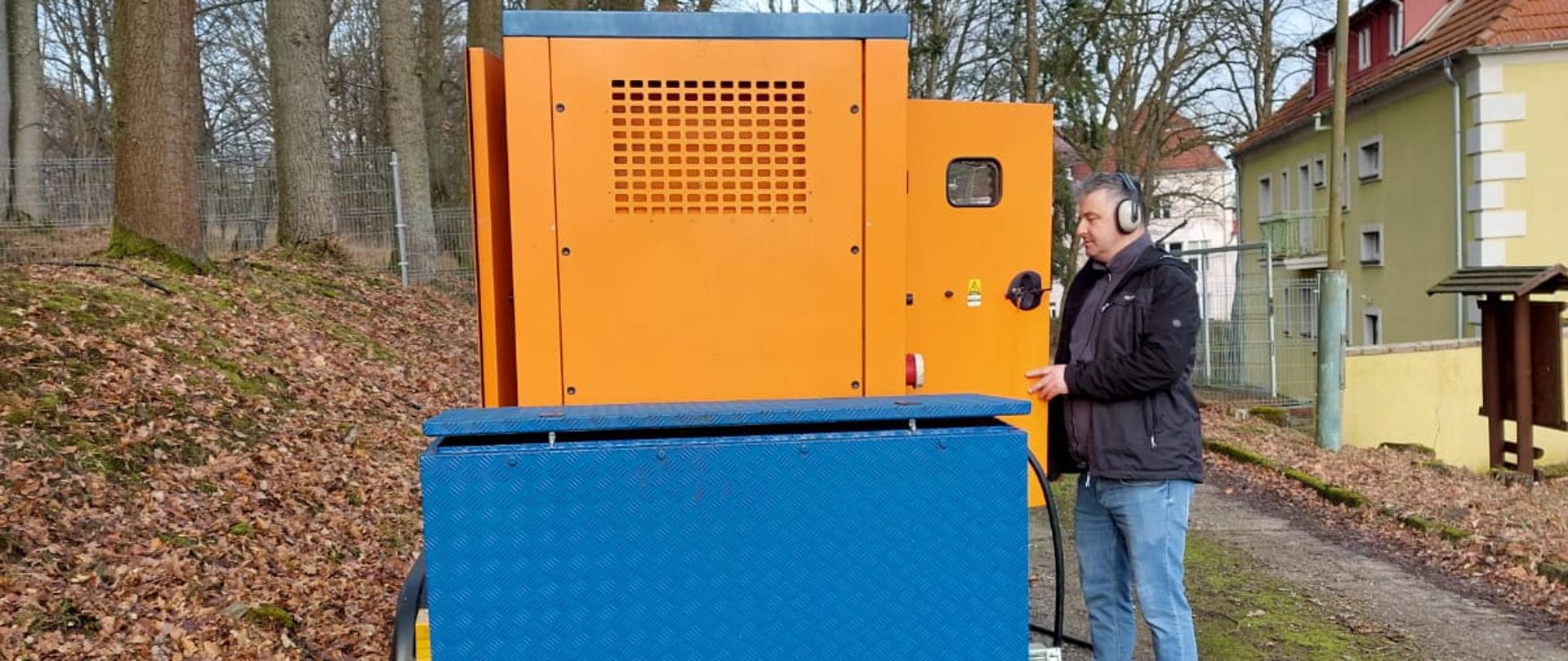 Zdjęcie przedstawia mobilny agregat prądotwórczy, który zasila uzdrowisko. Stoi on na drodze w tle drzewa. Obok agregatu znajduje się elektryk obsługujący go.