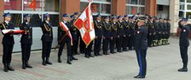 Zdjęcie zrobione na zewnątrz, na placu przed komendą powiatową Państwowej Straży Pożarnej podczas uroczystości. Generał Państwowej Straży Pożarnej salutuje sztandarowi Komendy Powiatowej Państwowej Straży Pożarnej.