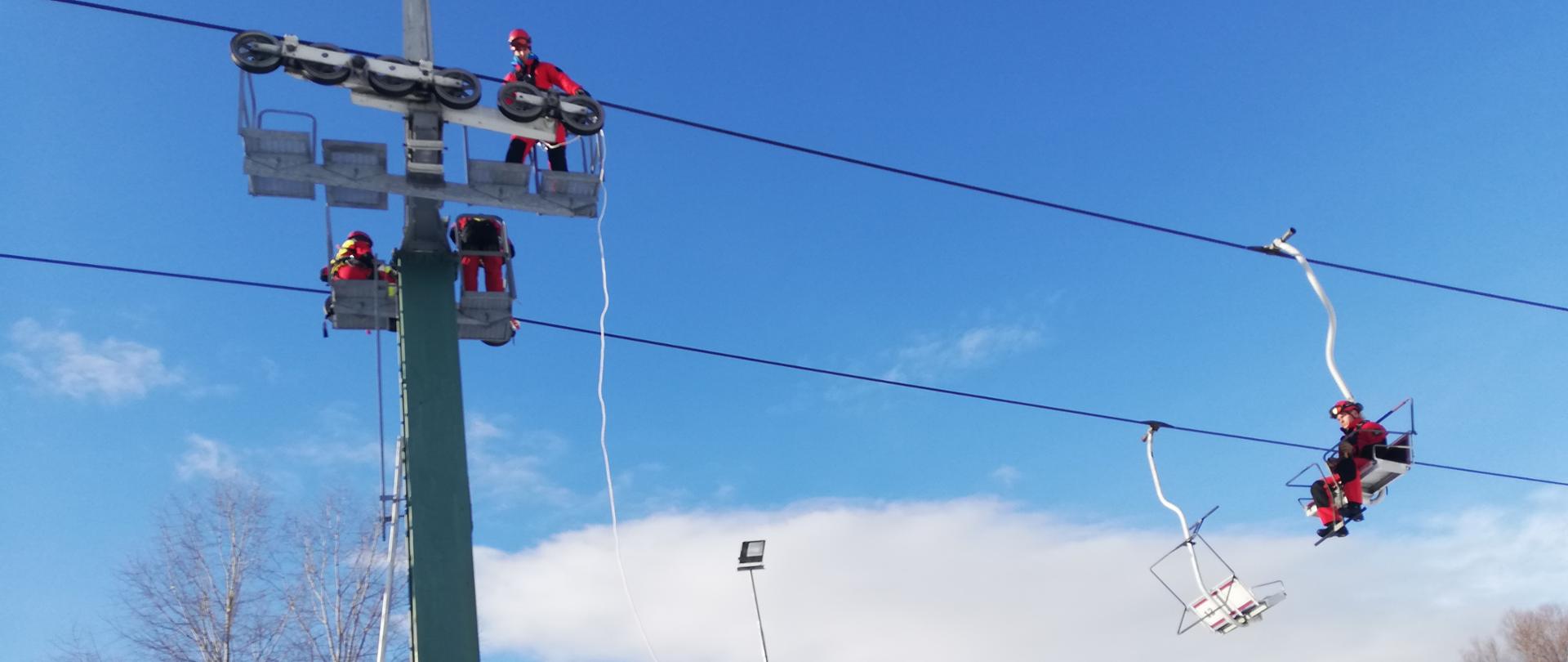 5 ratowników ze Specjalistycznej Grupy Ratownictwa Wysokościowego z Rzeszowa prowadzi ćwiczenia na wyciągu narciarskim. Zdjęcie ukazuje dwóch ratowników pozorujących osoby poszkodowane siedzących na dwóch krzesełkach, dwóch innych ratowników dociera do pozorantów przy użyciu technik i sprzętu ratownictwa wysokościowego celem ich ewakuacji. Piąty z ratowników po zjeździe znajduje się na zaśnieżonym podłożu.