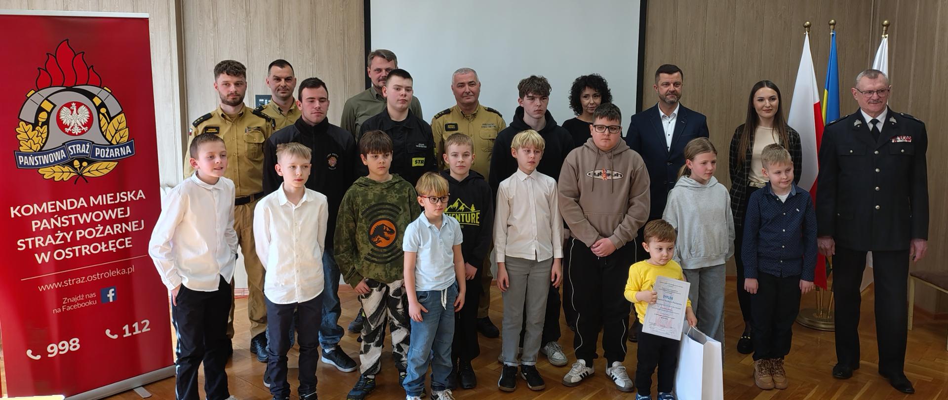 Zdjęcie grupowe uczestników i organizatorów eliminacji powiatowych Ogólnopolskiego Konkursu Wiedzy Pożarniczej „Młodzież zapobiega pożarom”. W centrum stoją laureaci i uczestnicy w różnym wieku, niektórzy trzymają dyplomy i upominki. Za nimi rzędem ustawili się przedstawiciele Państwowej Straży Pożarnej w piaskowych i ciemnych mundurach oraz osoby cywilne w strojach oficjalnych. Po lewej stronie widoczny jest duży czerwony baner Komendy Miejskiej PSP w Ostrołęce, a w tle biały ekran projekcyjny oraz polskie i unijne flagi. Całość dokumentuje oficjalne zakończenie etapu konkursu.