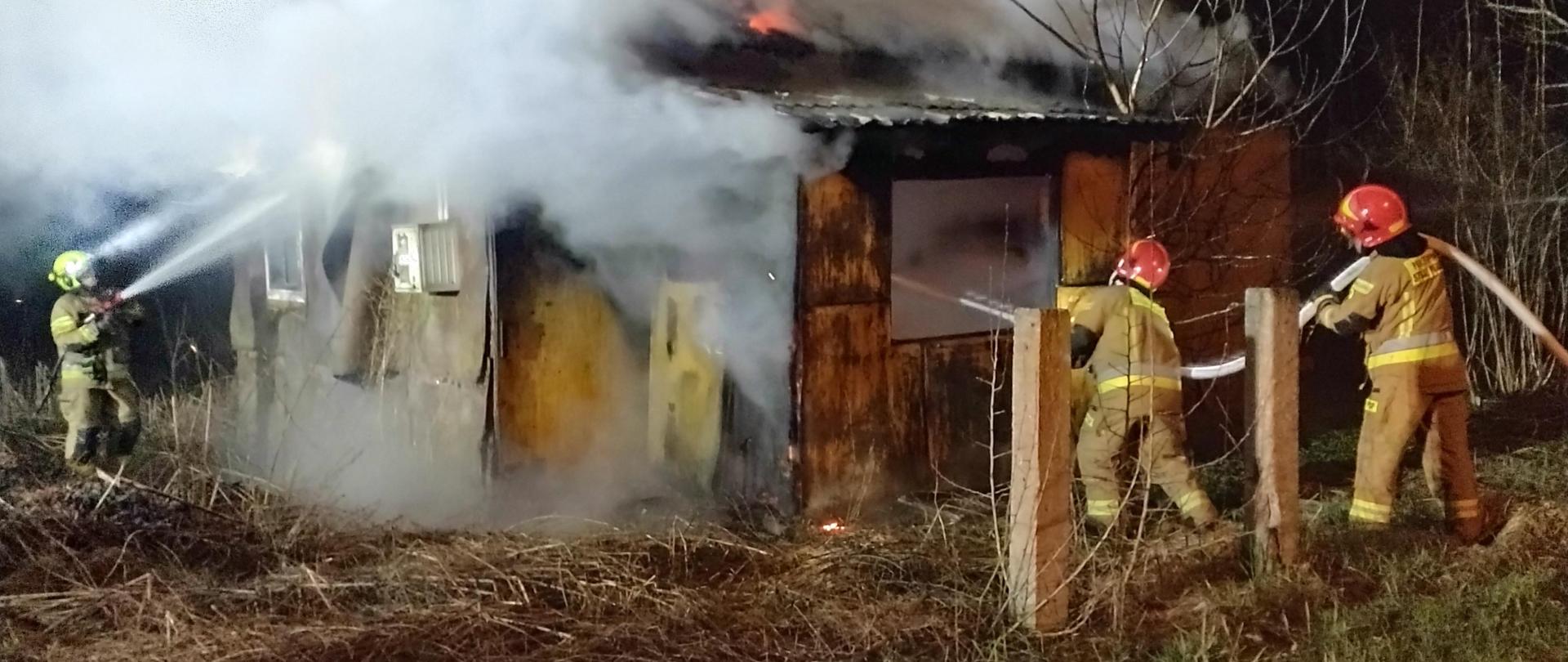 Płonący drewniany budynek, dym wydobywający się ze wszystkich otworów (okiennych, drzwiowych), trzech strażaków z liniami gaśniczymi, podaje wodę na pożar. 