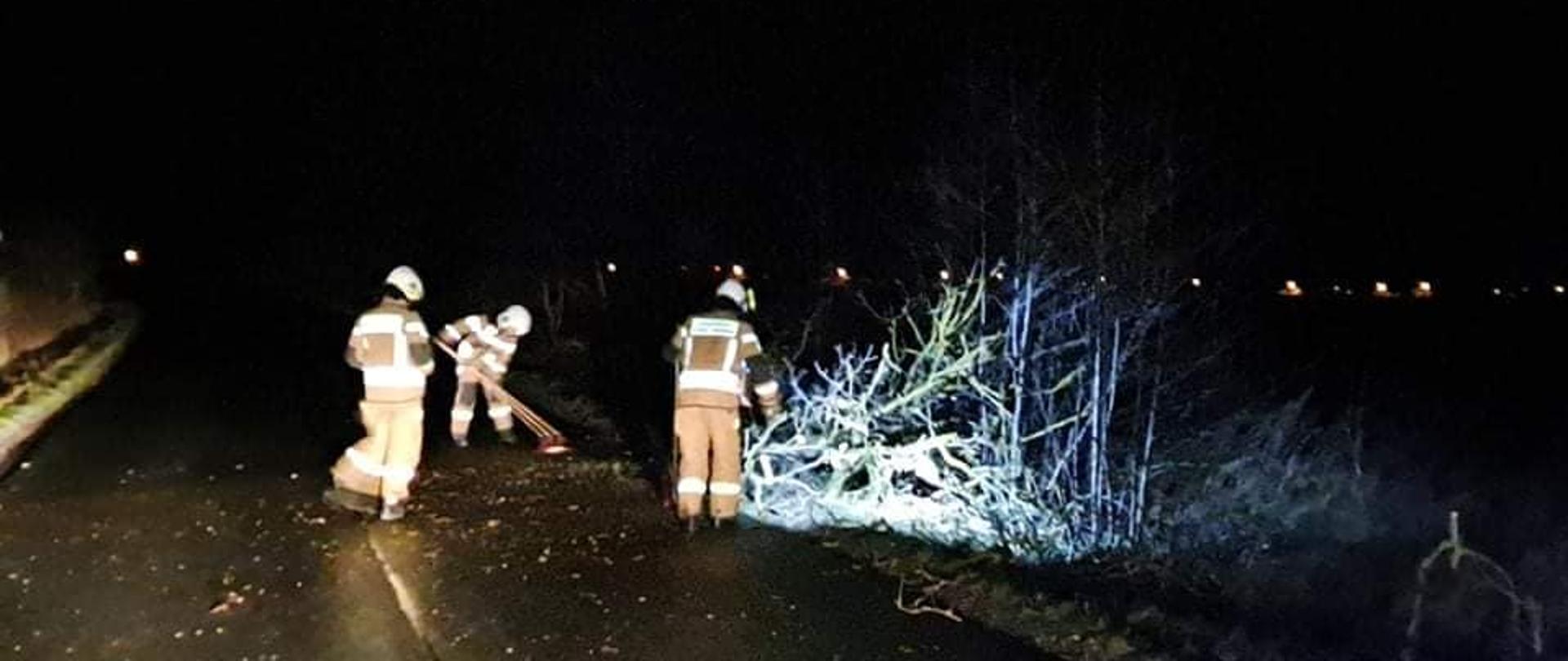 Zdjęcie zrobione w porze nocnej, strażacy usuwają połamane gałęzie z drogi, jeden z nich zmiata resztki konarów za pomocą szczoty