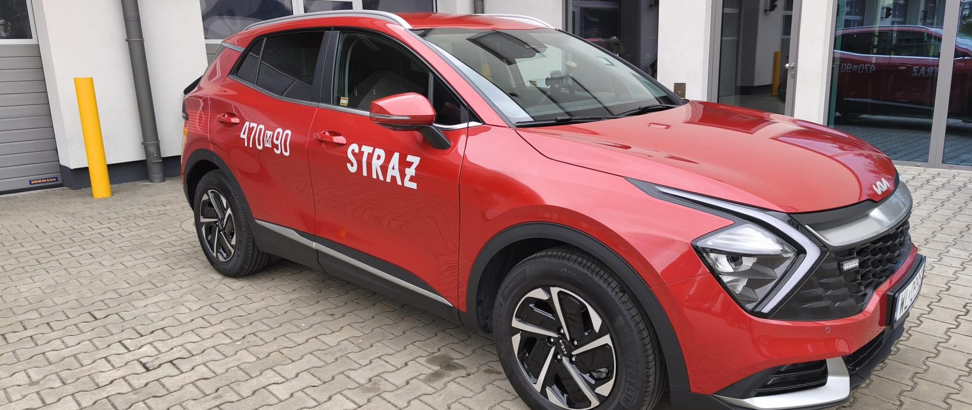 Na zdjęciu widać samochód osobowy w kolorze czerwony stojący na kostce brukowej. W tle widać bramy garażowe. Z boku pojazd posiada naklejkę z napisem Straż oraz z numerem 470m90. 