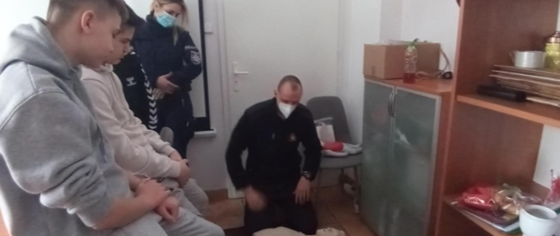 Strażak udzielający pokazu pierwszej pomocy. Strażak klęczy przy manekinie a obok stoją uczestnicy pogadanki.