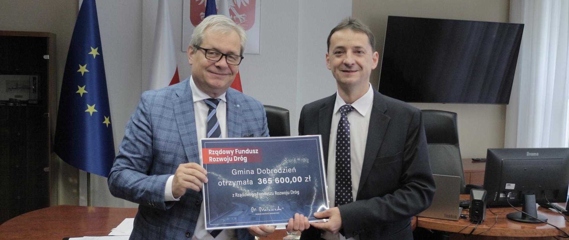 Na zdjęciu wojewoda opolski z burmistrzem Dobrodzienia. Stoją obok siebie, pozują do zdjęcia z wydrukiem czeku informującego o przyznanym dofinansowaniu. W tle biuro oraz biało-czerwone flagi.