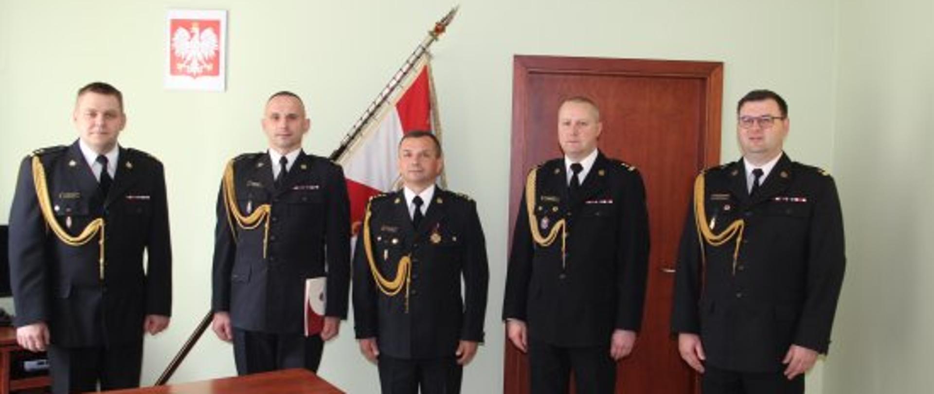 na tle jasnej ściany stoi pięciu mężczyzn strażaków ubranych w ciemne mundury ze złotymi sznurami, za nimi widać sztandar oraz godło 