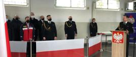 Na garażu JRG po lewej widać część trybuny honorowej, na której stoją zaproszeni goście. W środku zdjęcia przy mównicy z godłem RP stoi ustępujący Komendant Powiatowy PSP - przemawia do zgromadzonych. Strażacy w umundurowaniu wyjściowym. 