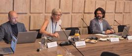 Za stołem prezydialnym siedzą trzy osoby, dwóch mężczyzn a pomiędzy nimi kobieta, która przemawia do mikrofonu. Na stołach widać włączone komputery, mikrofony i rozłożone notatki. W tle widać drewnopodobną ścianę z napisem Ministerstwo Klimatu i Środowiska. 