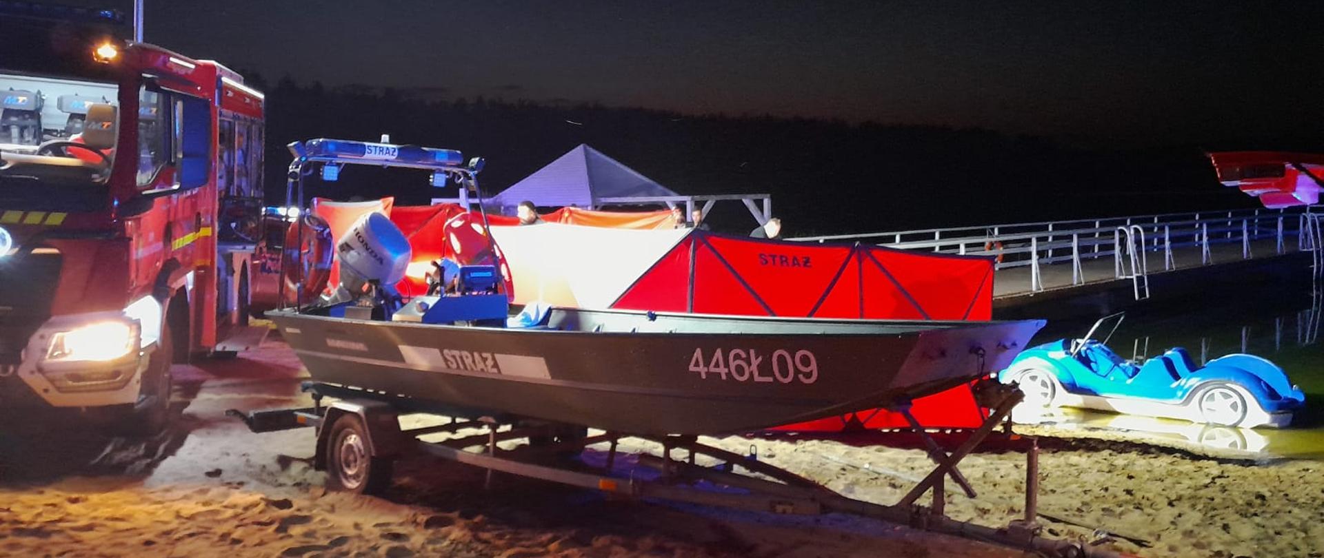 Łódź ratownicza za którym znajduje się ustawiony na brzegu jeziora parawana osłaniający miejsce udzielanej pomocy.