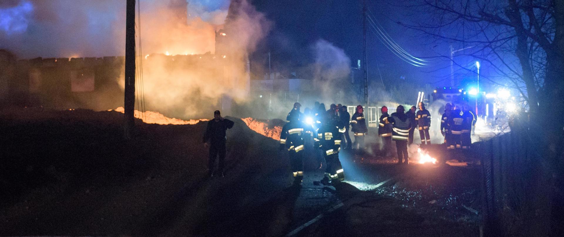 noc, duża grupa strażaków stoi na drodze, z boku widoczny pożar, 