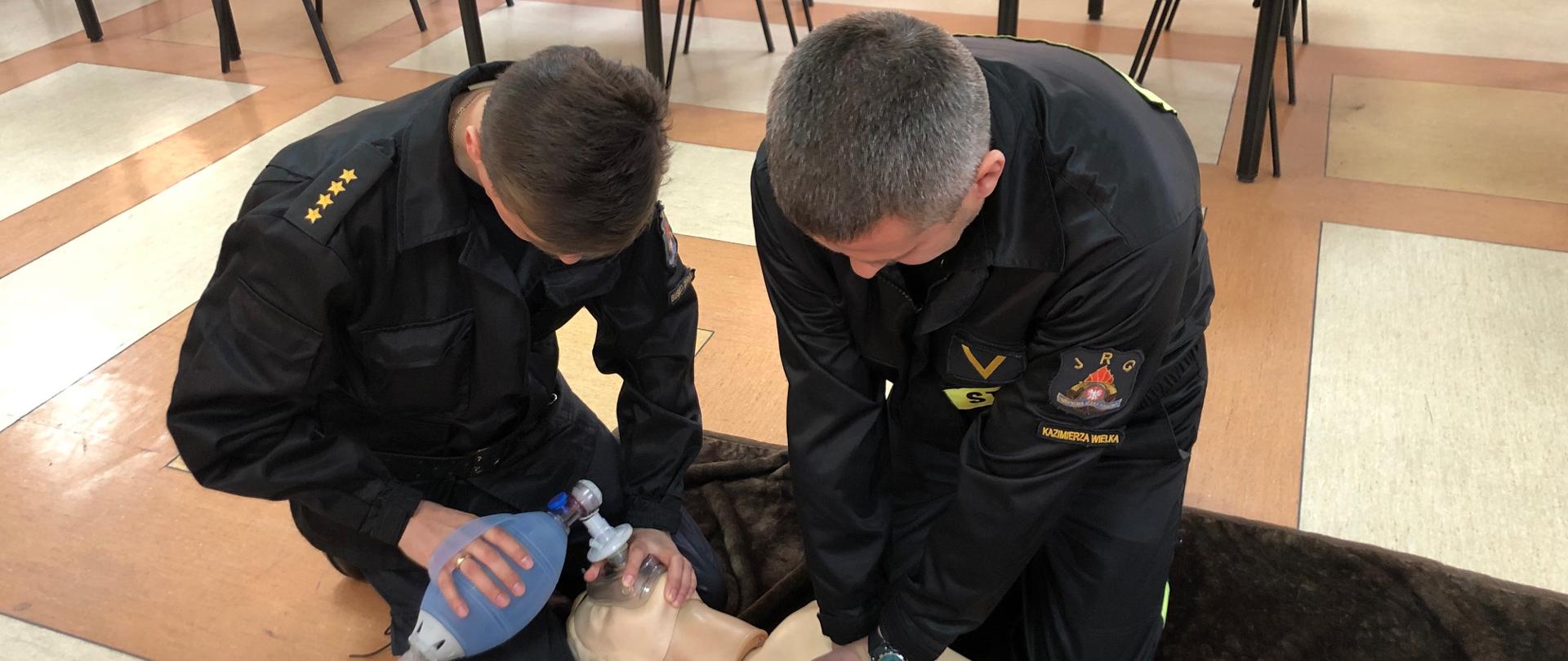 Na pierwszym zdjęciu strażacy-ratownicy wykonują symulację resuscytacji krążeniowo-oddechowej u osoby dorosłej. Jeden z ratowników wykonuje 30 uciśnięć klatki piersiowej, natomiast drugi wykonuje 2 oddechy ratownicze za pomocą worka samorozprężalnego.