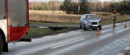 Z lewej strony tył samochodu ratowniczego stojący na jednym z pasów drogi. Po prawej uszkodzony samochód osobowy na pasie drogi oraz strażak stojący bokiem.