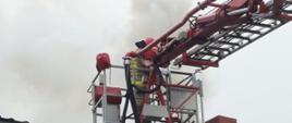 Pożar sadzy w kominie. Na pierwszym planie widoczny fragment budynku gospodarczego wykonanego z cegły. Nad dachem budynku znajduje się kosz podnośnika hydraulicznego, Znajdujący się w nim strażak gasi pożar sadzy w kominie.
