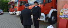 Zmiany kadrowe w rawickiej komendzie. Plac przed komendą powiatową, na którym odbywa się uroczysty apel. Na tle samochodów pożarniczych komendant powiatowy składa gratulacje strażakowi, który złożył ślubowanie. Z prawej strony roll-ap i stolik, na którym leży teczka. 