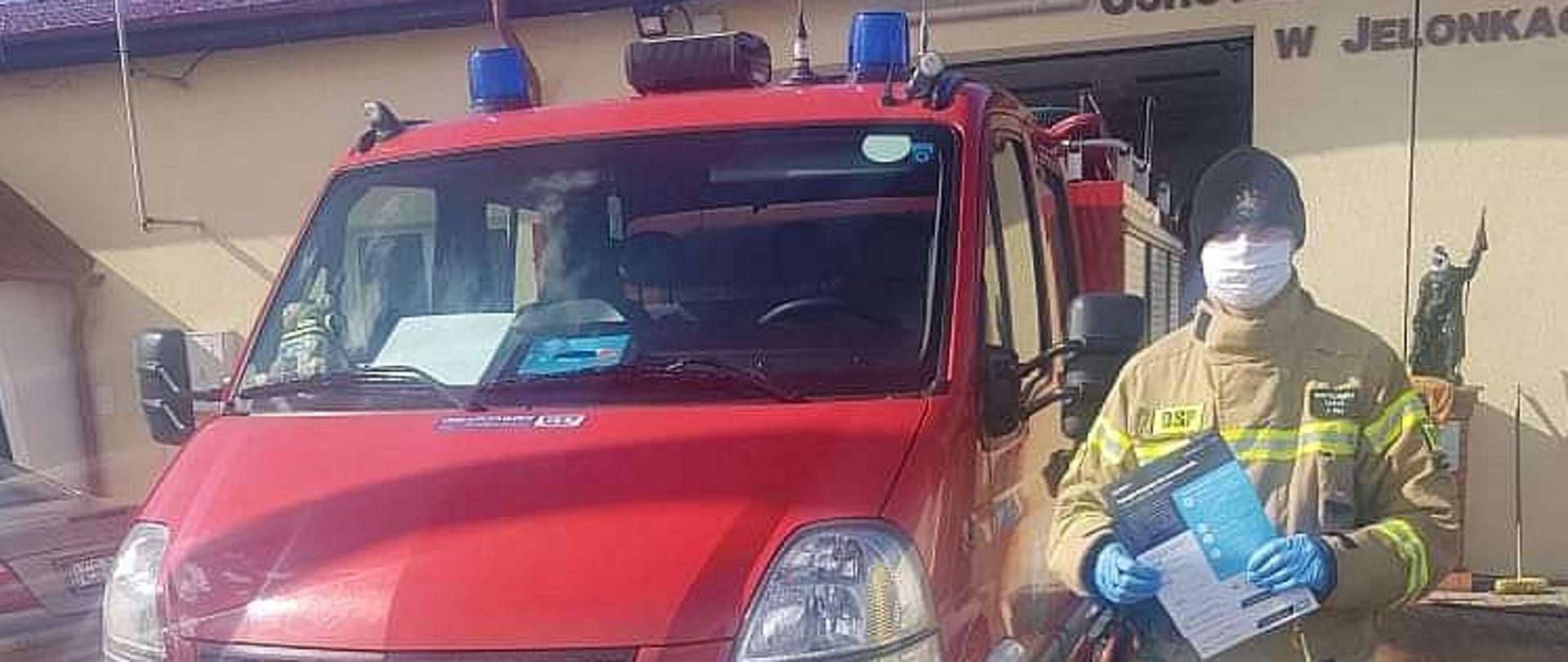 Strażak ochotnik stojący obok samochodu pożarniczego trzymający w ręku ulotkę dotyczącą szczepień. W tle budynek remizy ochotniczej straży pożarnej w Jelonkach.