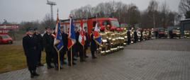 Zdjęcie przedstawia funkcjonariuszy Państwowej Straży Pożarnej ustawionych podczas uroczystego apelu połączonego z przekazaniem nowych samochodów pożarniczych. Od lewej ustawiony jest dowódca pocztów. Funkcjonariusz ubrany w płaszcz oraz mundur galowy. Na prawo od niego ustawieni w czterech rzędach po trzy osoby są funkcjonariusze tworzący poczty sztandarowe. Funkcjonariusze ubrani w kurtki służbowe oraz mundury galowe. Pierwszy z nich w każdym rzędzie w rękach trzyma sztandar. Dalej za nimi ustawieni są w dwu szeregu pozostali strażacy. Ubrani są oni w ubrania specjalne w kolorze piaskowym. Na głowach pozakładane mają hełmy. W oddali widać pojazdy pożarnicze, których to nastąpi przekazanie. Pojazdy w kolorze czerwonym z białymi i srebrnymi elementami zabudowy. 