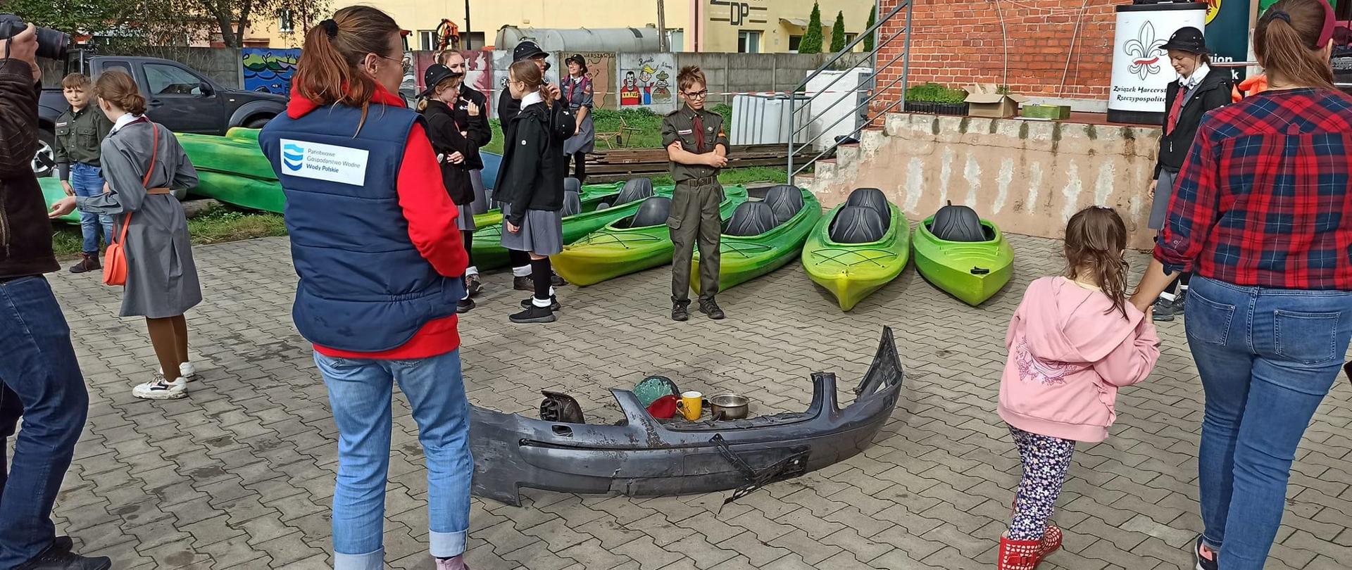 VIII Jesienne Sprzątanie Noteci. Na pierwszym planie pracownica Wód Polskich w kamizelce z logo. Stoi obok zebranych śmieci. W tle harcerze i kajaki leżące na betonie. Dookoła przechodnie. 