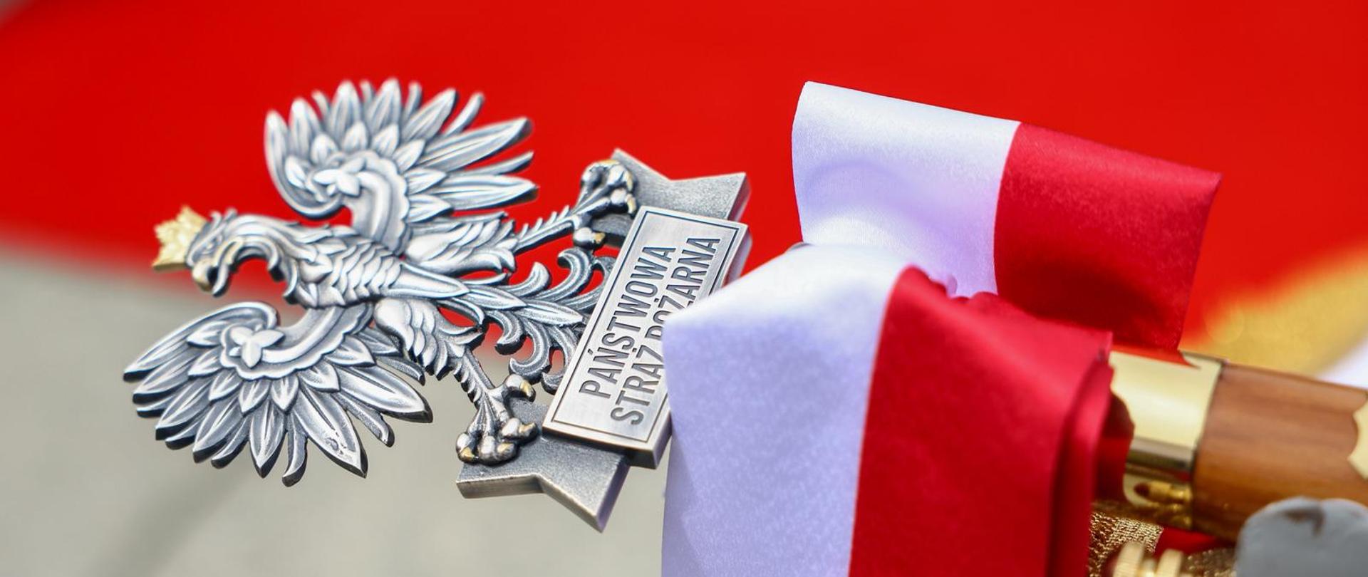 Zbliżenie na głowicę drzewca sztandaru w kształcie srebrnego orła z napisem „PAŃSTWOWA STRAŻ POŻARNA”. Do drzewca przywiązana jest biało-czerwona wstęga.