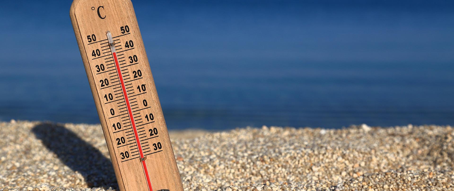 Termometr wskazujący wysoką temperaturę, ok 40'C wbity w żwirową plażę. W tle woda.