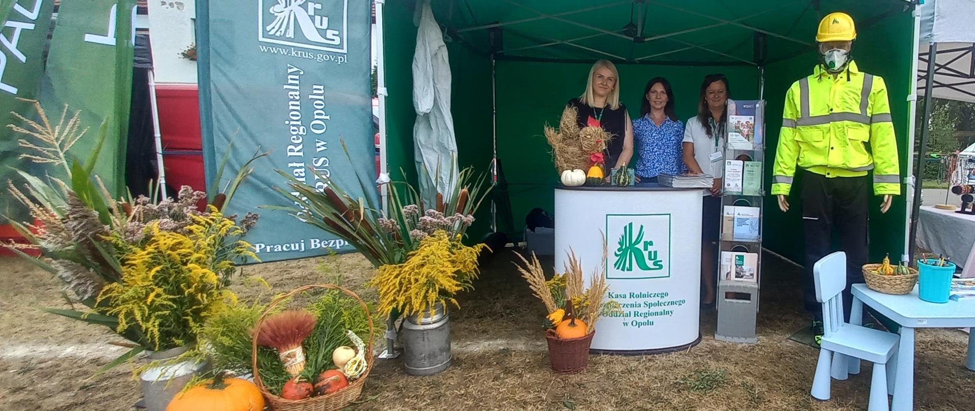 Trzy kobiety pozują przy stoisku pod zielonym namiotem z logo KRUS. Po lewej stronie dożynkowe dekoracje z ziół, traw i warzyw. Po prawej stoi manekin prezentujący odzież ochronną.