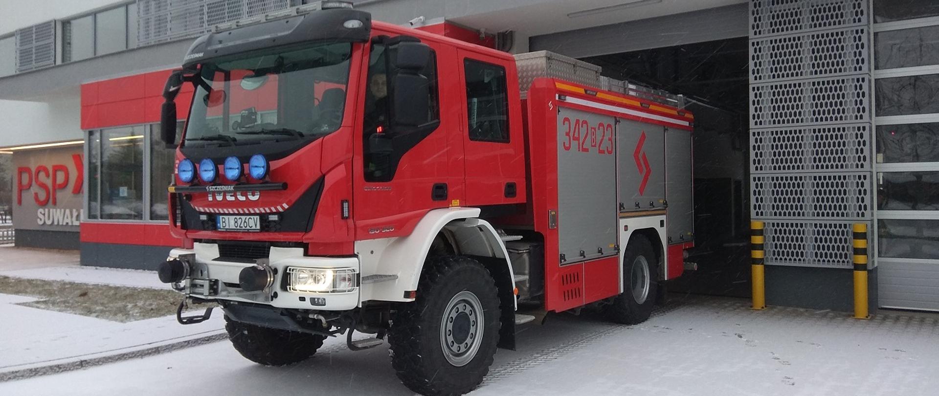Samochód średni ratowniczo gaśniczy w kolorze czerwonym. W dalszej części widać część budynku komendy z napisem PSP w Suwałkach