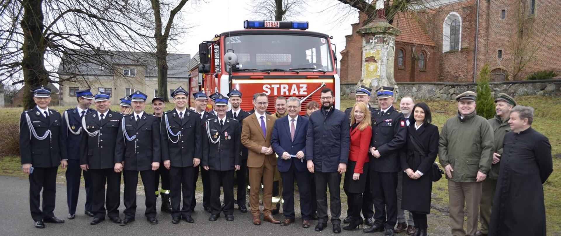 Na zdjęciu znajdują się uczestnicy przekazania samochodu ratowniczo gaśniczego, w tle kościół, samochód ratowniczo gaśniczy, budynek mieszkalny i drzewa.