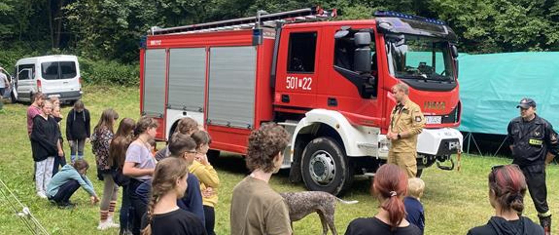 Na zdjęciu widzimy grupę ludzi stojących w lesie na terenie obozu harcerskiego. Widać też Strażacki pojazd ratowniczy oraz strażaków udzielających instruktażu harcerzom. .