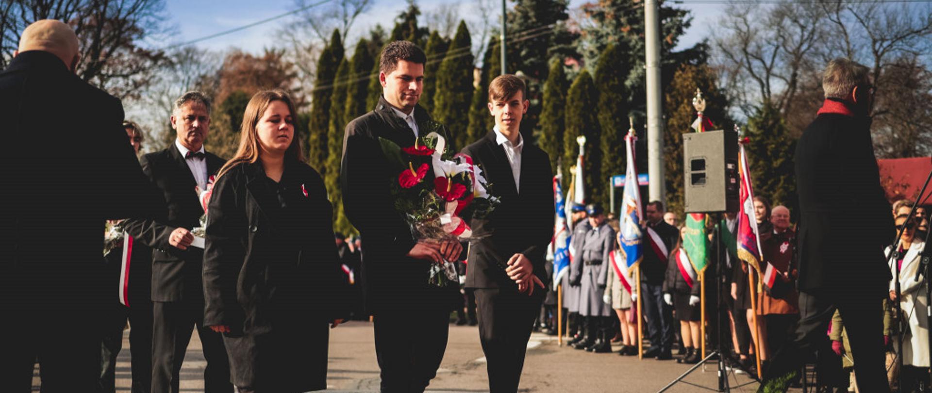 zdjęcie przedstawia członków Samorządu Uczniowskiego i jego opiekuna w trzyosobowej delegacji z bukietem biało-czerwonych kwiatów w ręku podczas składanie kwiatów pod pomnikiem wolności w mielcu, w tle widać poczty sztandarowe, drzewa i bezchmurne niebo