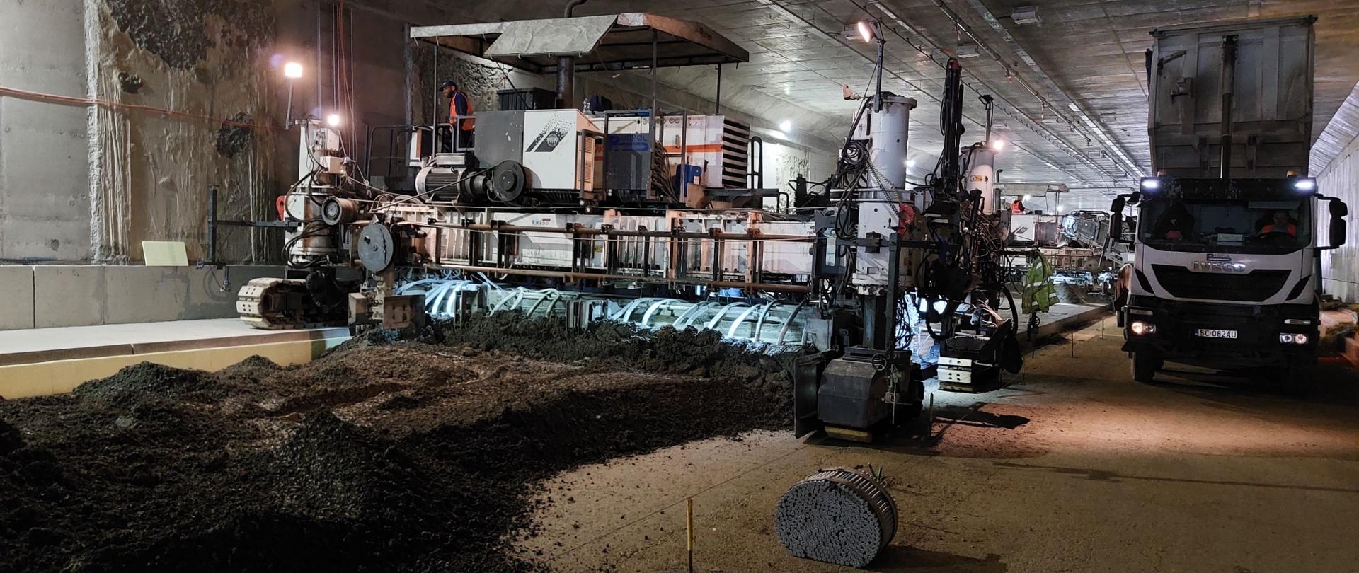 tunel drogowy, po lewej stronie pracuje maszyna układająca nawierzchnię betonową, po prawej ciężarówka