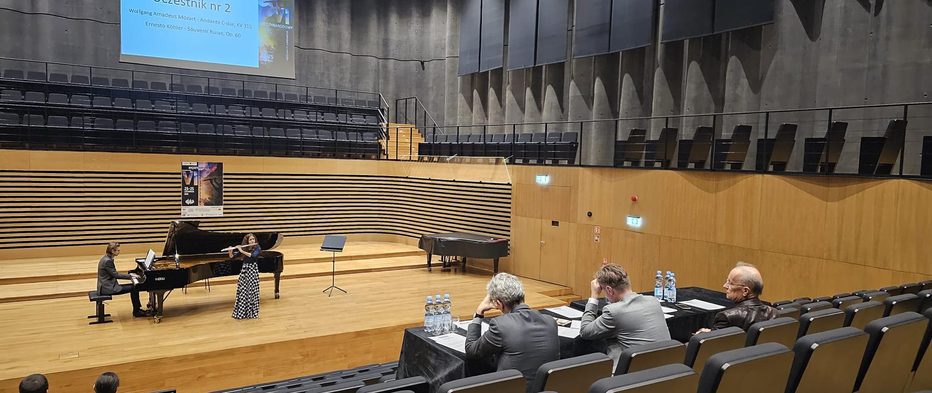 Nastolatka stojąc na środku estrady sali koncertowej PSM gra na flecie poprzecznym, za nią na fortepianie gra mężczyzna. Z przodu widać od tyłu widownię oraz siedzące przy stole Jury.