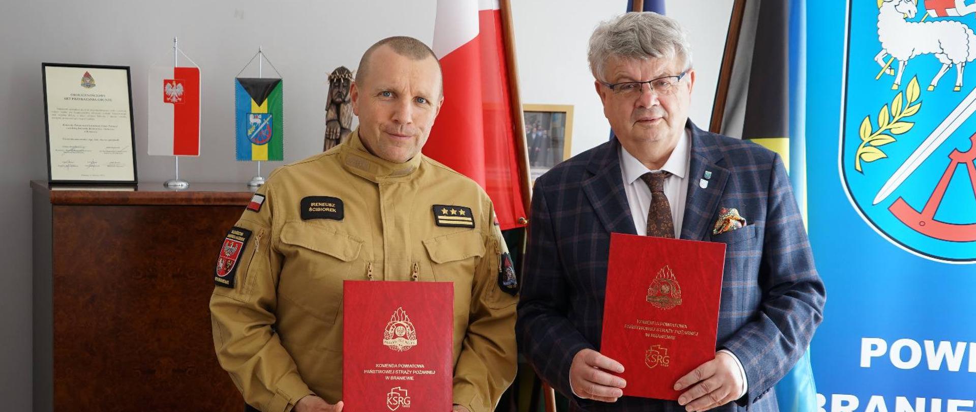 Na zdjęciu widzimy dwóch mężczyzn stojących obok siebie, trzymających w rękach czerwone teczki z emblematem straży pożarnej oraz napisem "Krajowy System Ratowniczo-Gaśniczy".
Po lewej stronie znajduje się strażak w mundurze koloru piaskowego. Po prawej stronie stoi elegancko ubrany mężczyzna w marynarce w kratę i krawacie, starosta braniewski.
W tle znajdują się flagi (w tym Polski), herb powiatu braniewskiego oraz dokument w ramce, zdjęcie zostało wykonane podczas oficjalnego podpisania porozumienia i współpracy pomiędzy strażą pożarną a samorządem powiatowym. Atmosfera jest oficjalna i uroczysta.
