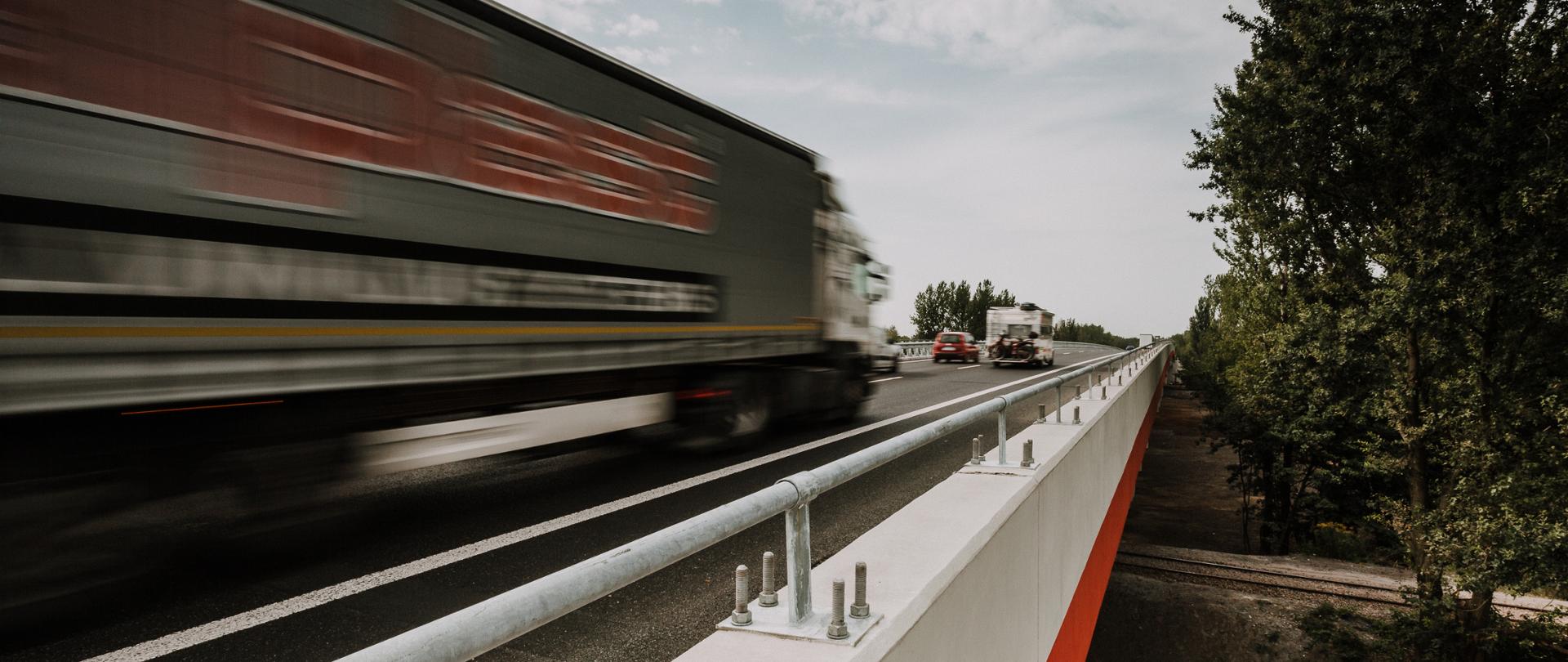 Fotografia drogi z poruszającymi się po niej samochodami. Na pierwszym planie widoczny pojazd ciężarowy (TIR), przed nim kamper i pojazd osobowy.
W lewej części fotografii widoczna zieleń przydrogowa. 