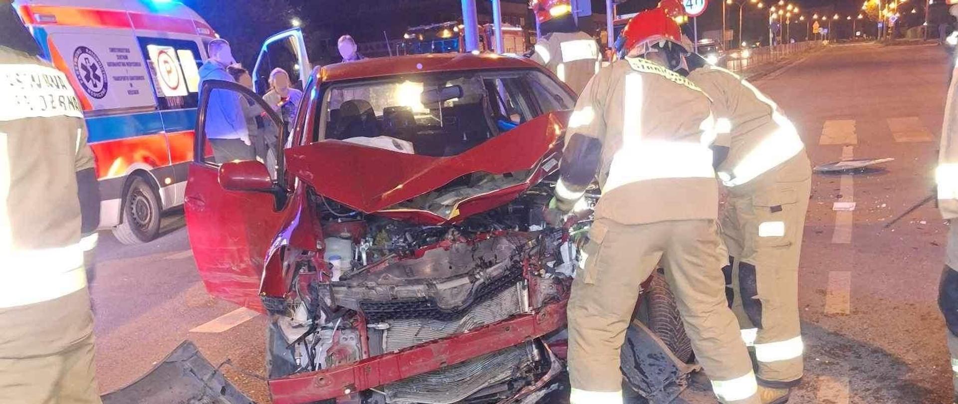 Na pierwszym planie samochód osobowy z uszkodzonym przodem pojazdu, przy nim dwóch strażaków odłączających akumulator, z tyłu karetka pogotowia.