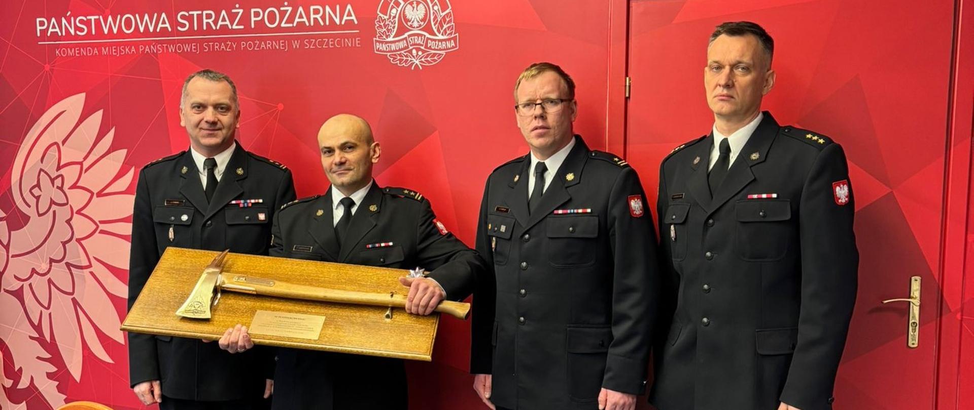 Fotografia przedstawia czterech mężczyzn w galowych mundurach Państwowej Straży Pożarnej, stojących w rzędzie we wnętrzu pomieszczenia z czerwonymi drzwiami i logo PSP na ścianie. Mężczyzna drugi od lewej trzyma drewnianą deskę z zamontowanym na niej toporkiem strażackim, przypuszczalnie upominek lub odznaczenie. Wszyscy mężczyźni patrzą w kierunku obiektywu.