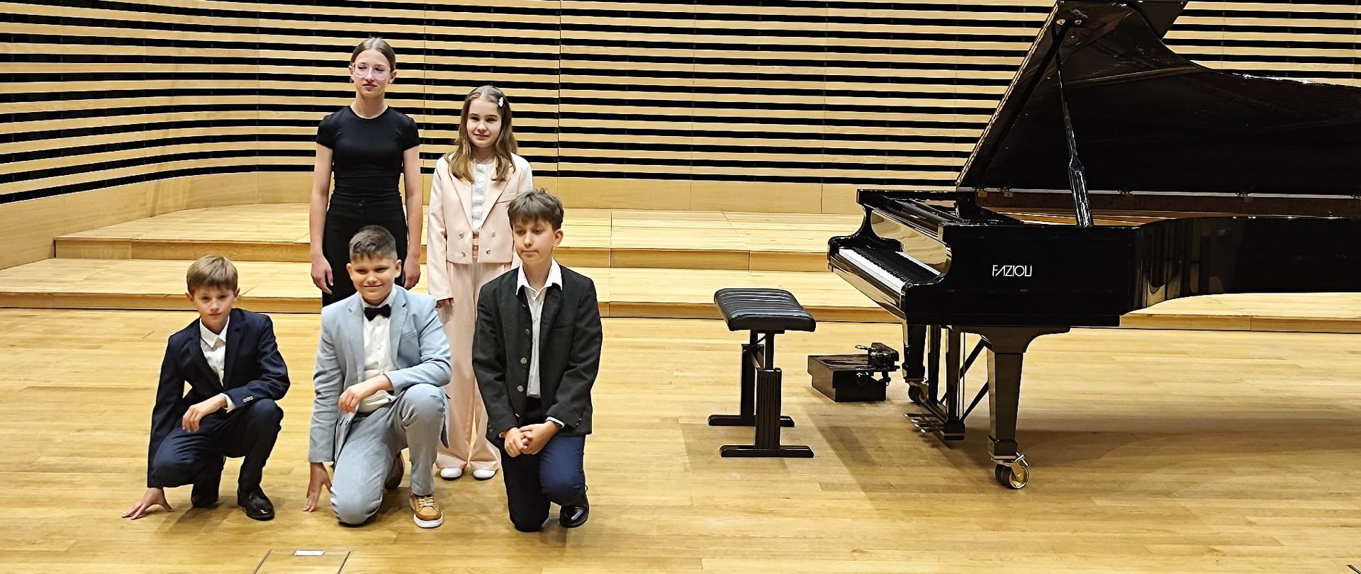 Dwie dziewczynki i trzech chłopców stoją na scenie przy fortepianie.